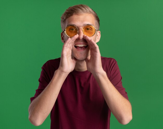 Chico guapo joven alegre con camisa roja y gafas llamando a alguien aislado en la pared verde