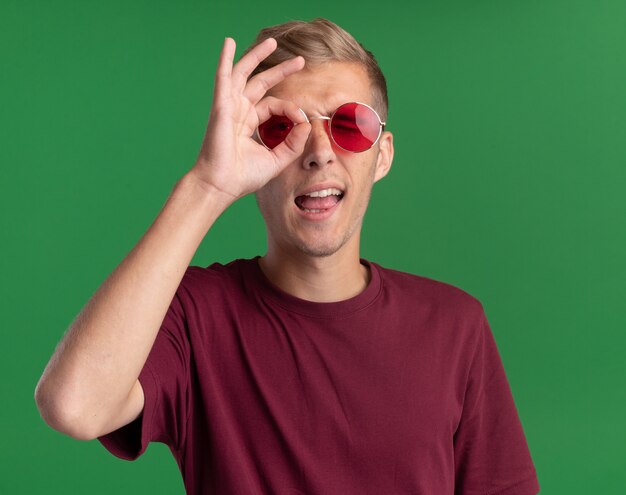 Chico guapo joven alegre con camisa roja y gafas mostrando gesto de mirada aislado en la pared verde