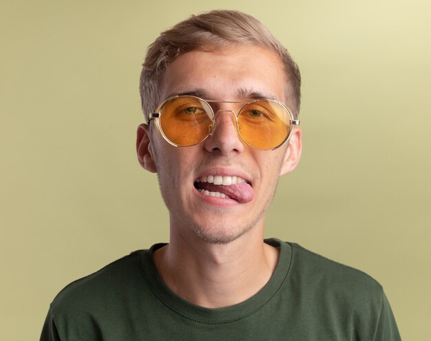 Chico guapo joven alegre con camisa verde con gafas mostrando la lengua aislada en la pared verde oliva