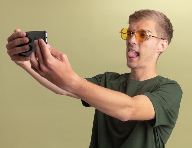 Chico guapo joven alegre con camisa verde con gafas mostrando la lengua y tomar un selfie aislado en la pared verde oliva