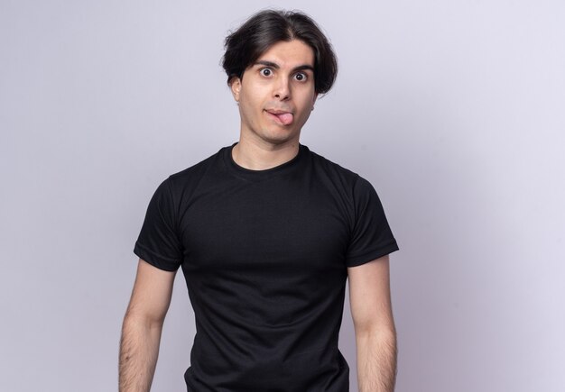 Chico guapo joven alegre con camiseta negra que muestra la lengua aislada en la pared blanca