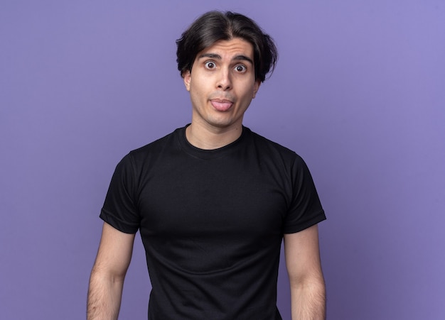 Chico guapo joven alegre con camiseta negra que muestra la lengua aislada en la pared púrpura