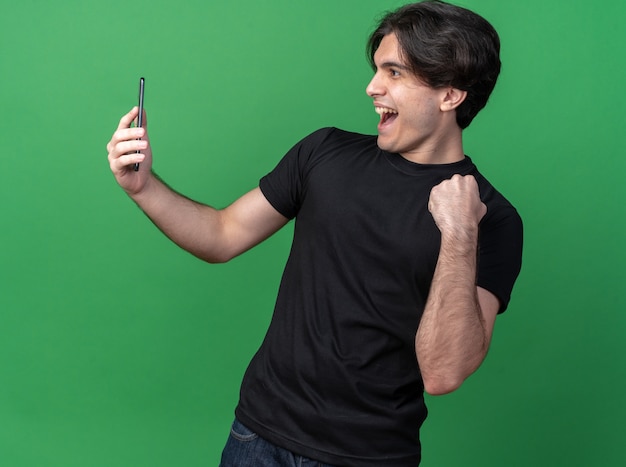 Chico guapo joven alegre con camiseta negra sosteniendo y mirando el teléfono mostrando sí gesto aislado en la pared verde