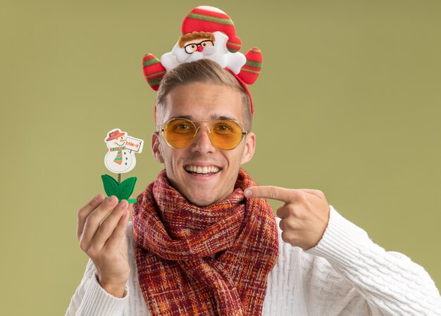 Chico guapo joven alegre con diadema y bufanda de santa claus sosteniendo y apuntando al juguete de muñeco de nieve aislado en la pared verde oliva