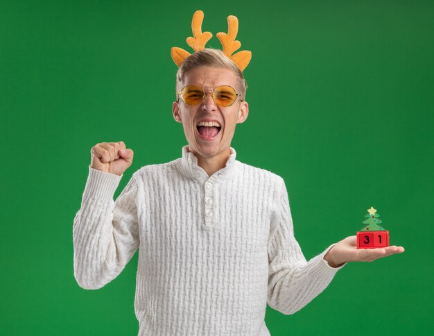 Chico guapo joven alegre con diadema de cuernos de reno con gafas sosteniendo el juguete del árbol de navidad con fecha haciendo gesto sí aislado en la pared verde