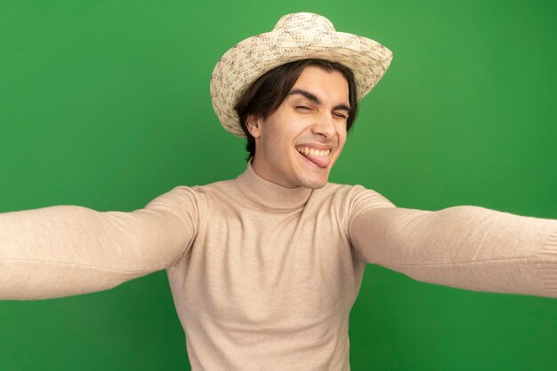 Chico guapo joven alegre con sombrero mostrando la lengua sosteniendo el frente aislado en la pared verde