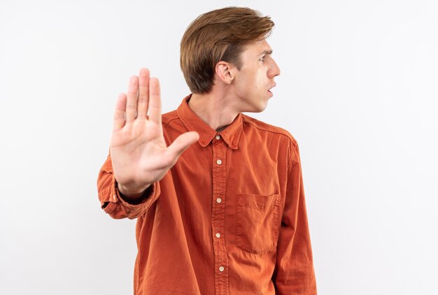 Chico guapo joven de aspecto confuso con camisa roja mostrando gesto de parada aislado en la pared blanca