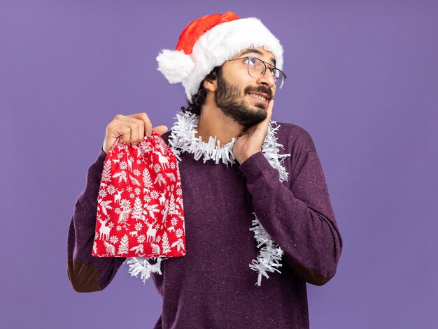 Chico guapo joven de aspecto impresionado con sombrero de navidad con guirnalda en el cuello sosteniendo una bolsa de navidad poniendo la mano en la mejilla aislada en la pared azul