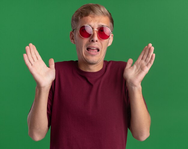 Chico guapo joven asustado con camisa roja y gafas extendiendo las manos aisladas en la pared verde