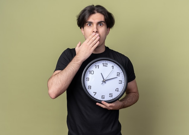 Chico guapo joven asustado con camiseta negra que sostiene el reloj de pared y la boca cubierta con la mano aislada en la pared verde oliva