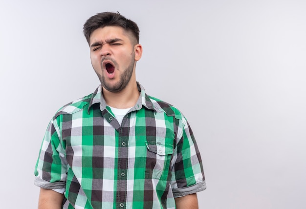 Chico guapo joven con camisa a cuadros bostezando con sueño cerrando los ojos de pie sobre la pared blanca