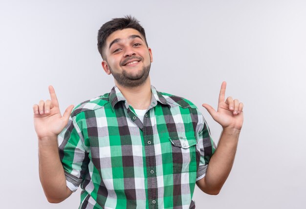 Chico guapo joven con camisa a cuadros felizmente mirando hacia arriba con los dedos índices de pie sobre la pared blanca