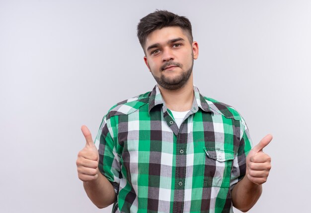 Chico guapo joven con camisa a cuadros haciendo feliz pulgares arriba de pie sobre la pared blanca