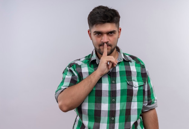 Chico guapo joven con camisa a cuadros haciendo señal de silencio con el dedo índice de pie sobre la pared blanca