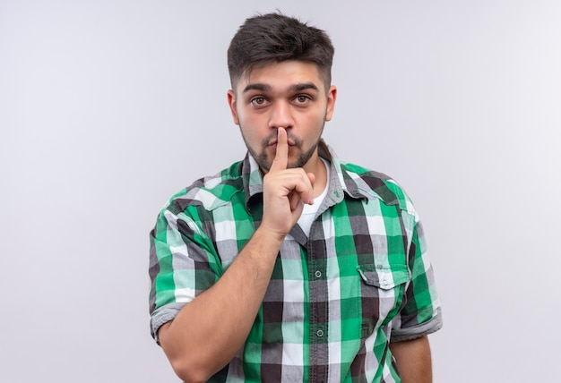 Chico guapo joven con camisa a cuadros haciendo señal de silencio de pie sobre la pared blanca