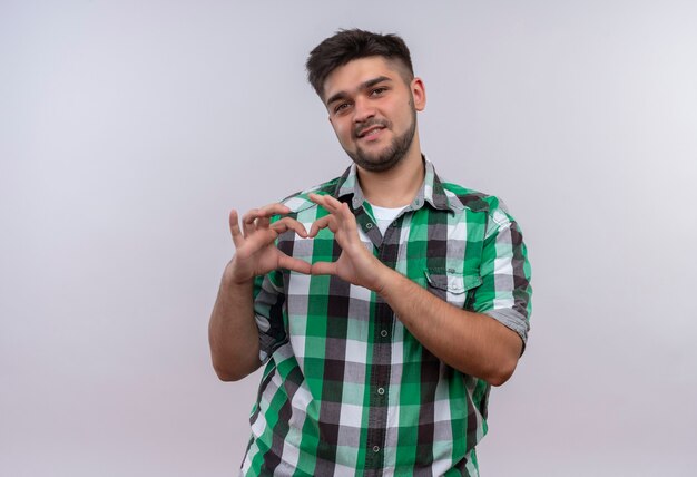 Chico guapo joven con camisa a cuadros haciendo signo de amor con las manos de pie sobre la pared blanca