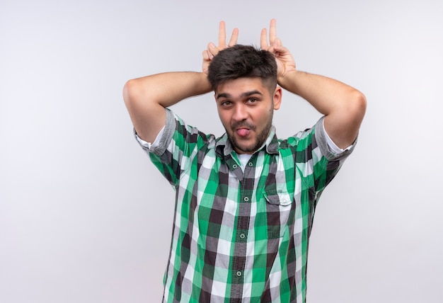 Foto gratuita chico guapo joven con camisa a cuadros juguetonamente haciendo cuernos y mostrando la lengua de pie sobre la pared blanca