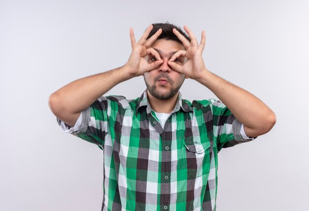 Chico guapo joven con camisa a cuadros juguetonamente haciendo gafas con los dedos de pie sobre la pared blanca