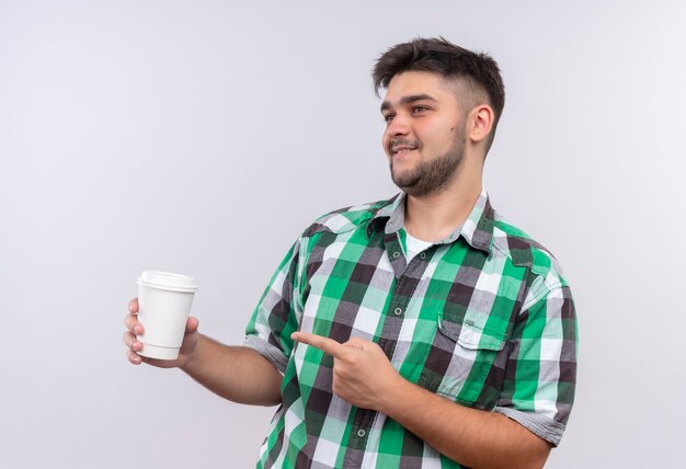 Foto gratuita chico guapo joven con camisa a cuadros mirando además complacido apuntando a la taza de café plastik de pie sobre la pared blanca