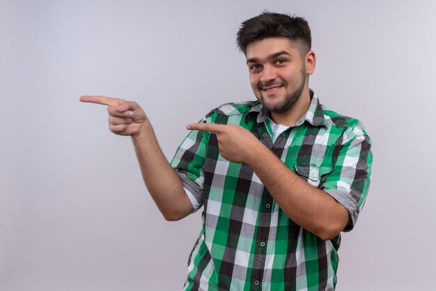 Chico guapo joven con camisa a cuadros mirando felizmente apuntando hacia la derecha con los dedos índices de pie sobre la pared blanca