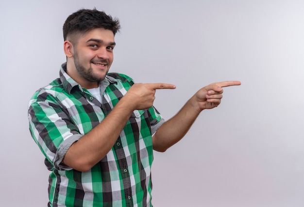 Chico guapo joven con camisa a cuadros mirando felizmente apuntando a la izquierda con los dedos índices de pie sobre la pared blanca