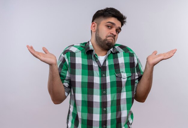 Chico guapo joven con camisa a cuadros mirando indeciso de pie sobre la pared blanca