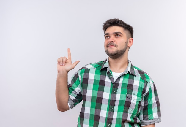 Chico guapo joven con camisa a cuadros mirando pensativamente hacia arriba haciendo el dedo índice de pie sobre la pared blanca