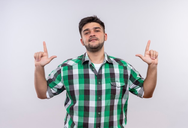 Chico guapo joven con camisa a cuadros mirando seriamente apuntando hacia arriba con los dedos índices de pie sobre la pared blanca