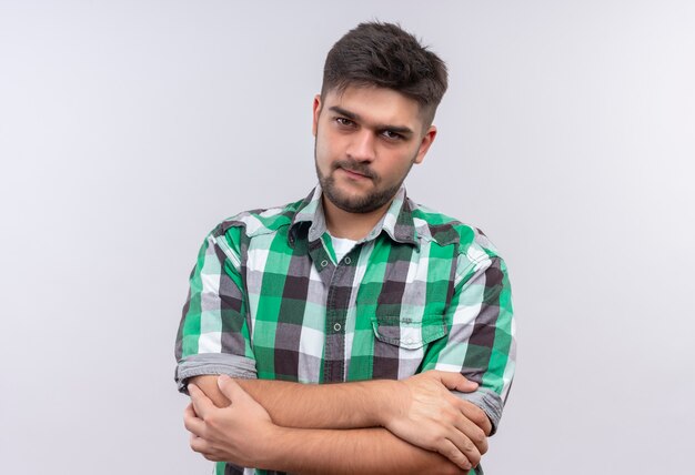 Chico guapo joven con camisa a cuadros mirando seriamente de pie sobre la pared blanca