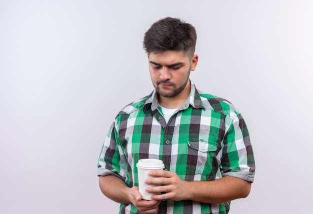 Chico guapo joven con camisa a cuadros mirando hacia su taza de café de plástico de pie sobre la pared blanca