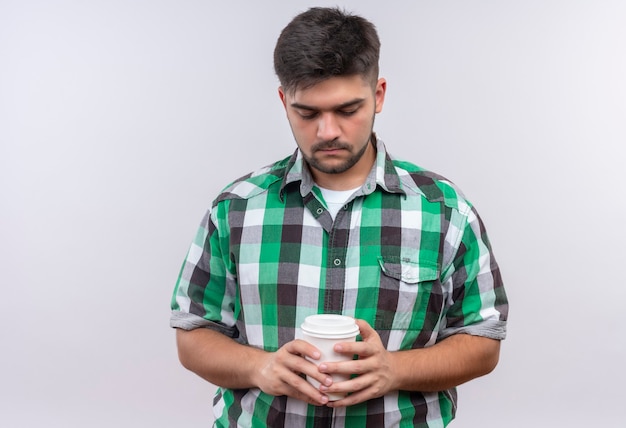 Chico guapo joven con camisa a cuadros mirando tristemente hacia abajo en su taza de café de plástico de pie sobre la pared blanca