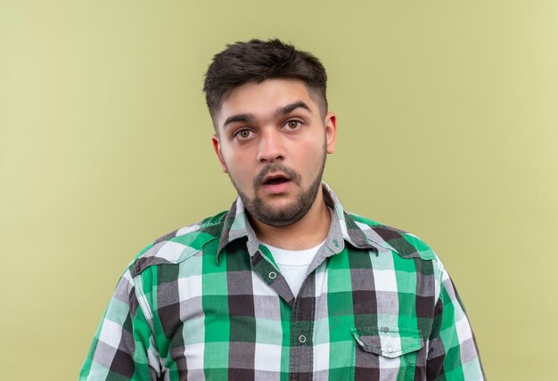 Chico guapo joven con camisa a cuadros de pie estupefacto sobre la pared de color caqui