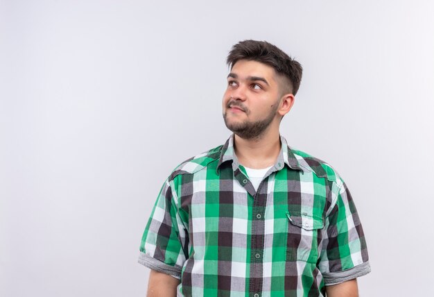 Chico guapo joven con camisa a cuadros de pie pensativamente sobre la pared blanca