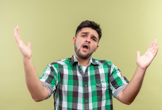 Chico guapo joven con camisa a cuadros que parece confundido no sabe qué hacer parado sobre la pared de color caqui