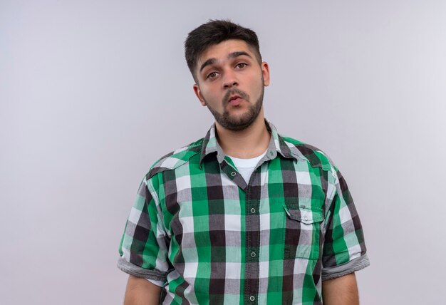 Chico guapo joven con camisa a cuadros silbidos de pie sobre la pared blanca