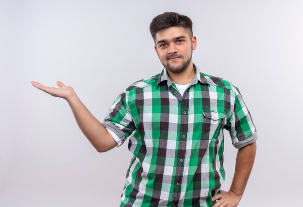 Chico guapo joven con camisa a cuadros sonriendo apuntando hacia la derecha con la mano de pie sobre la pared blanca