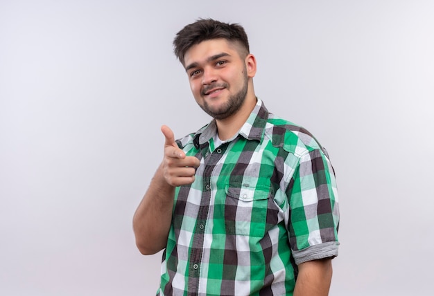 Foto gratuita chico guapo joven con camisa a cuadros sonriendo apuntando de pie sobre la pared blanca