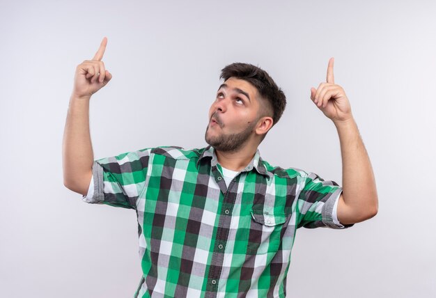 Chico guapo joven con camisa a cuadros sorprendido apuntando hacia arriba con el dedo índice de pie sobre la pared blanca