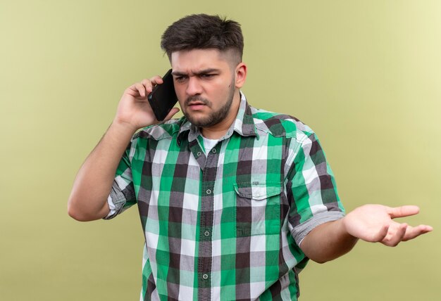 Chico guapo joven con camisa a cuadros sorprendido hablando por teléfono parado sobre la pared de color caqui