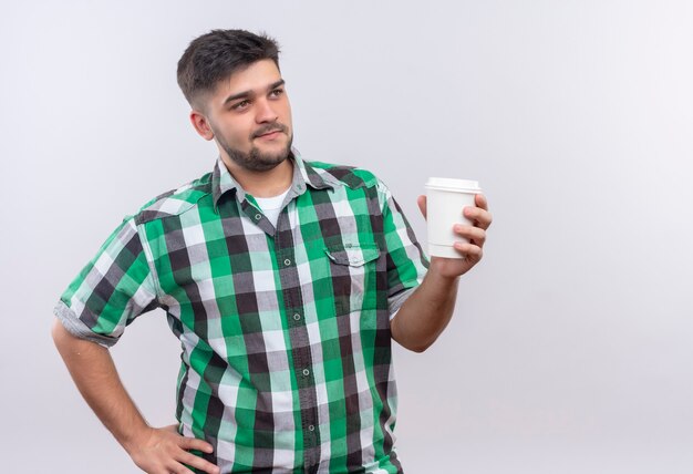 Chico guapo joven con camisa a cuadros sosteniendo una taza de café de plástico mirando además de estar parado sobre la pared blanca