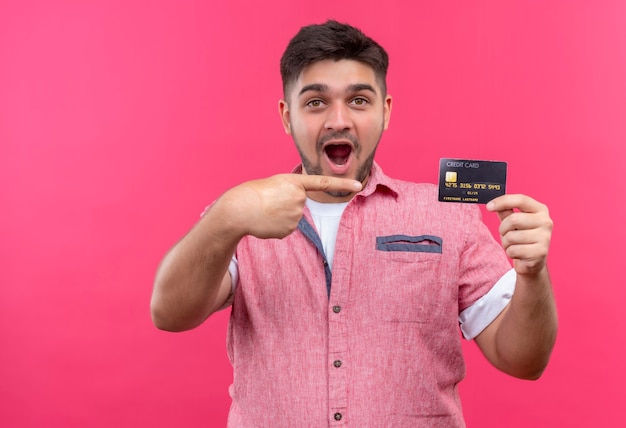 Chico guapo joven con camisa polo rosa apuntando a la tarjeta de pie sorprendido sobre la pared rosa