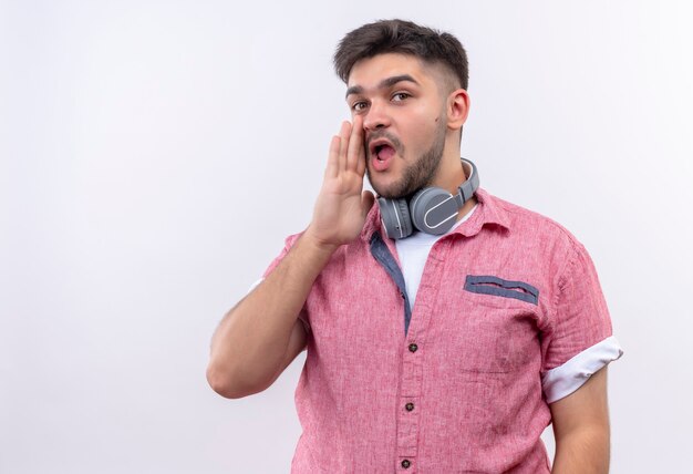 Chico guapo joven con camisa polo rosa y auriculares susurrando de pie sobre la pared blanca