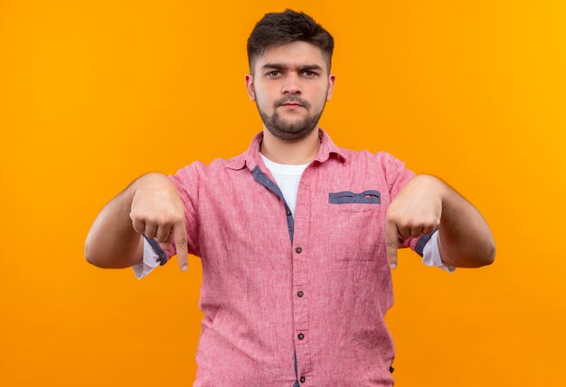 Chico guapo joven con camisa polo rosa disgustado mirando y haciendo el dedo índice hacia abajo de pie sobre la pared naranja