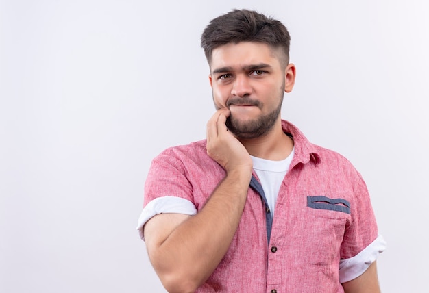 Chico guapo joven con camisa polo rosa dudosamente mirando de pie sobre la pared blanca