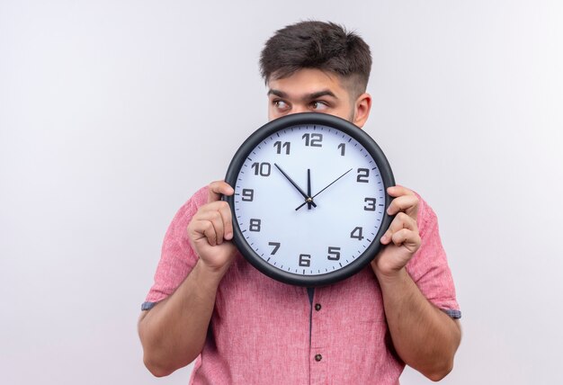 Foto gratuita chico guapo joven con camisa polo rosa escondido además del reloj preguntándose si está llegando tarde de pie sobre la pared blanca