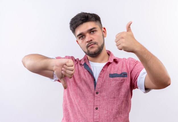 Chico guapo joven con camisa polo rosa indeciso mirando shwoing pulgares felices e infelices de pie sobre la pared blanca