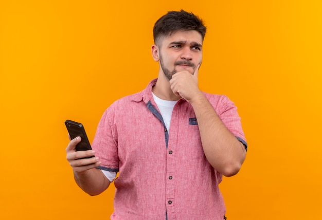 Chico guapo joven con camisa polo rosa mirando además de sostener cuidadosamente el teléfono de pie sobre la pared naranja