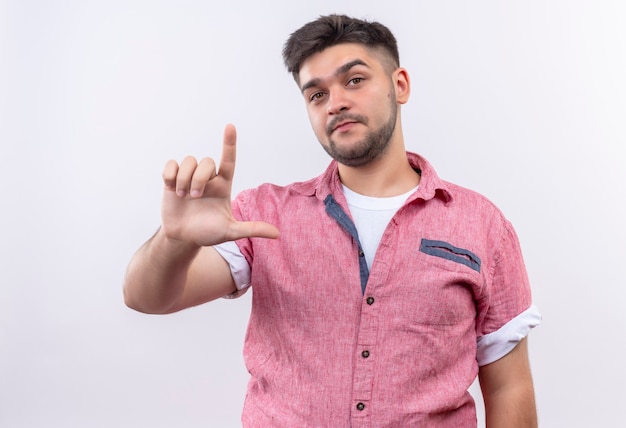 Chico guapo joven con camisa polo rosa mirando seriamente mostrando un signo más suelto con la mano de pie sobre la pared blanca