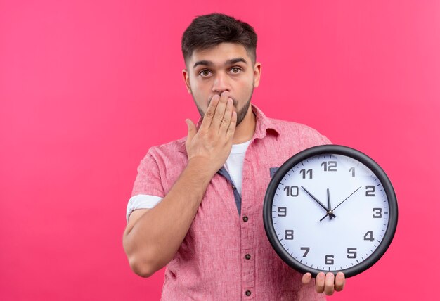 Chico guapo joven con camisa polo rosa mirando sorprendido sosteniendo el reloj con miedo de llegar tarde de pie sobre la pared rosa