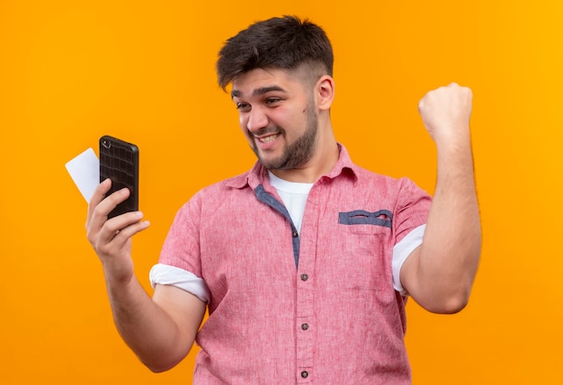 Chico guapo joven con camisa de polo rosa mirando el teléfono y la tarjeta regocijándose en la victoria de pie sobre la pared naranja